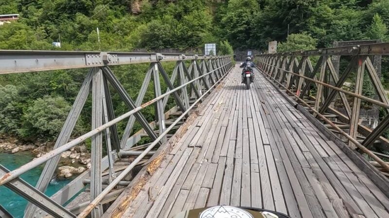 Rugged wooden bridge with metal support trusses and a motorcycle crossing over a lush green landscape.